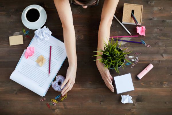 Above view of an unrecognizable woman sorting items on a working table, including stationery, documents, food, and a plant